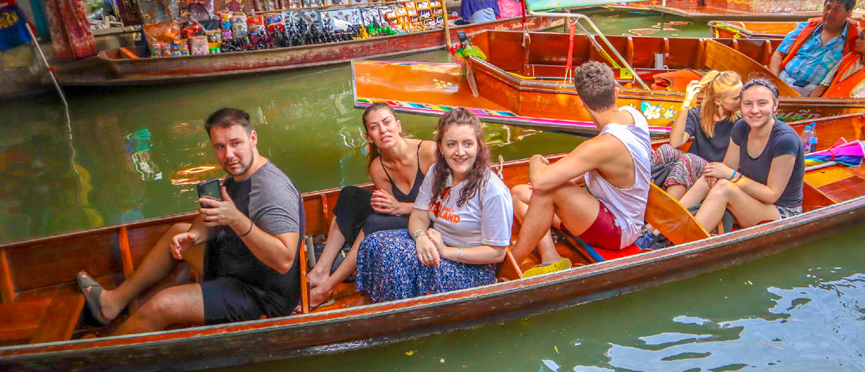 Happy tourists enjoying a boat ride through the bustling Amphawa Floating Market in Thailand.