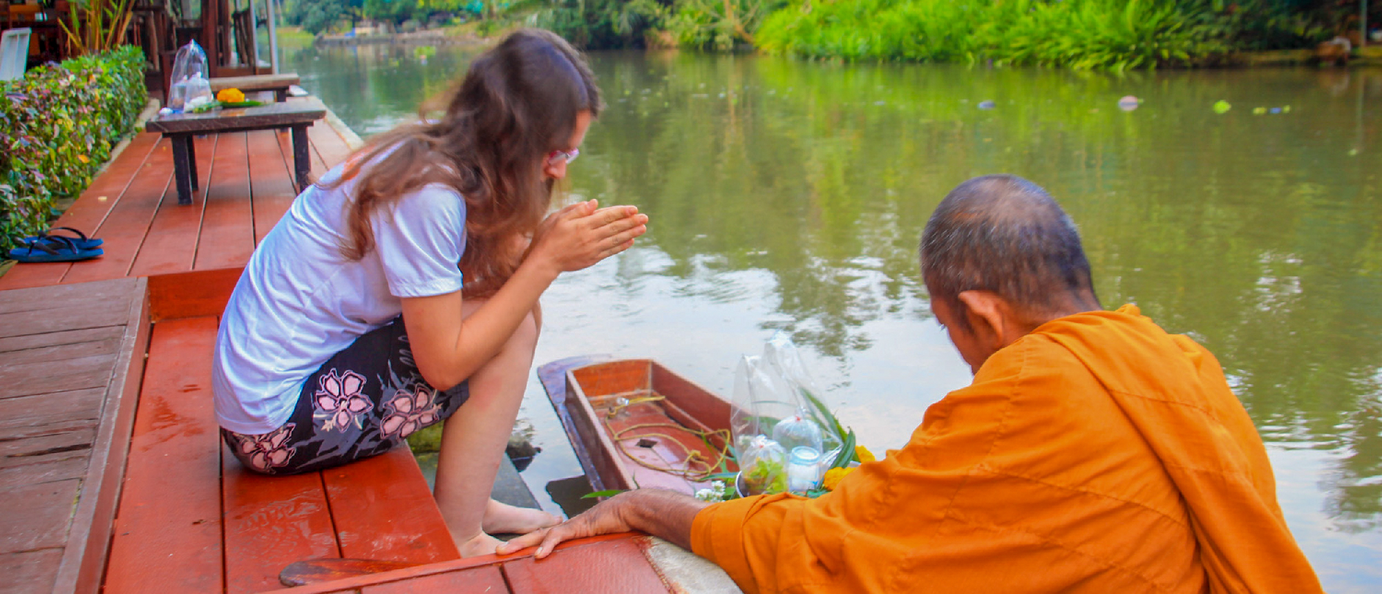 A woman offers alms to a monk from a wooden deck by a canal in Amphawa, Thailand.
