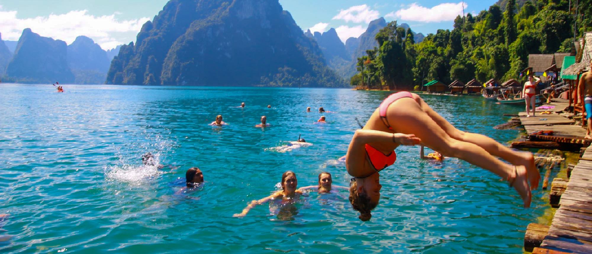 A young woman dives into the clear blue Cheow Lan Lake, with towering limestone karsts and bungalows.