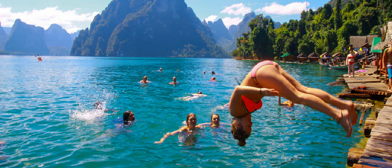 A young woman dives into the clear blue Cheow Lan Lake, with towering limestone karsts and bungalows.