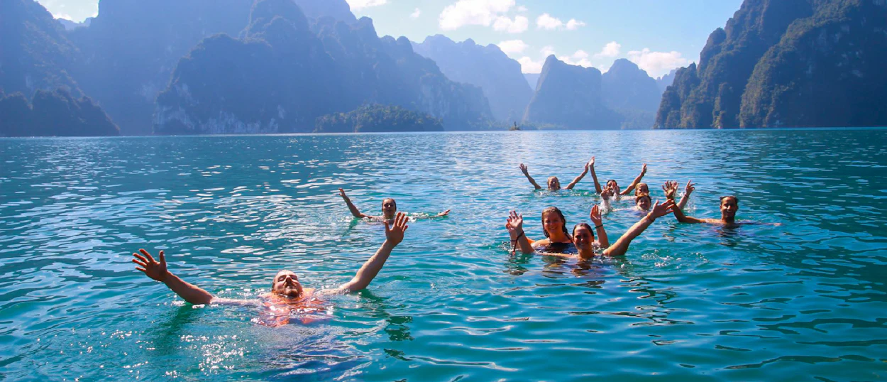 Excited backpackers swim in the clear blue waters of Cheow Lan Lake, surrounded by towering limestone mountains.