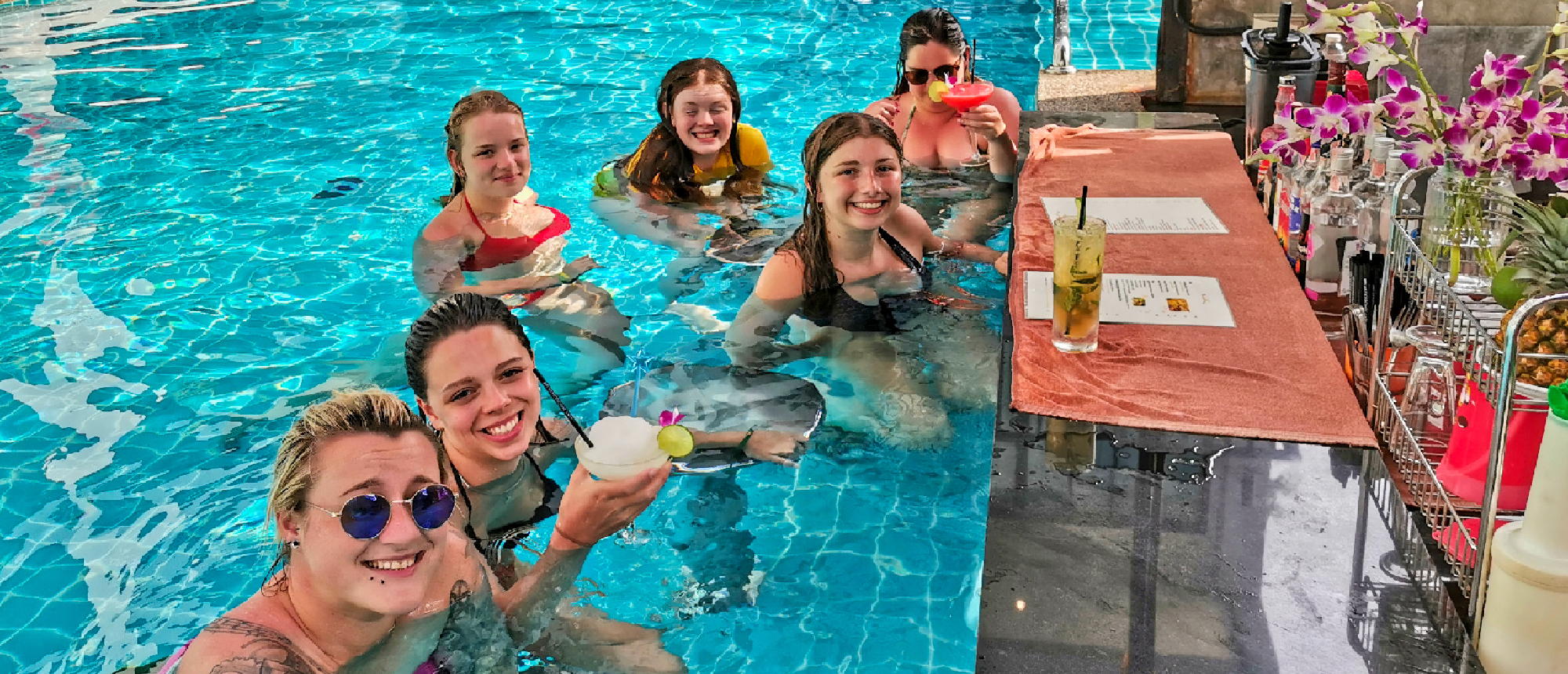 Six happy women enjoy drinks at a swim-up bar in a pool in Krabi, Thailand.