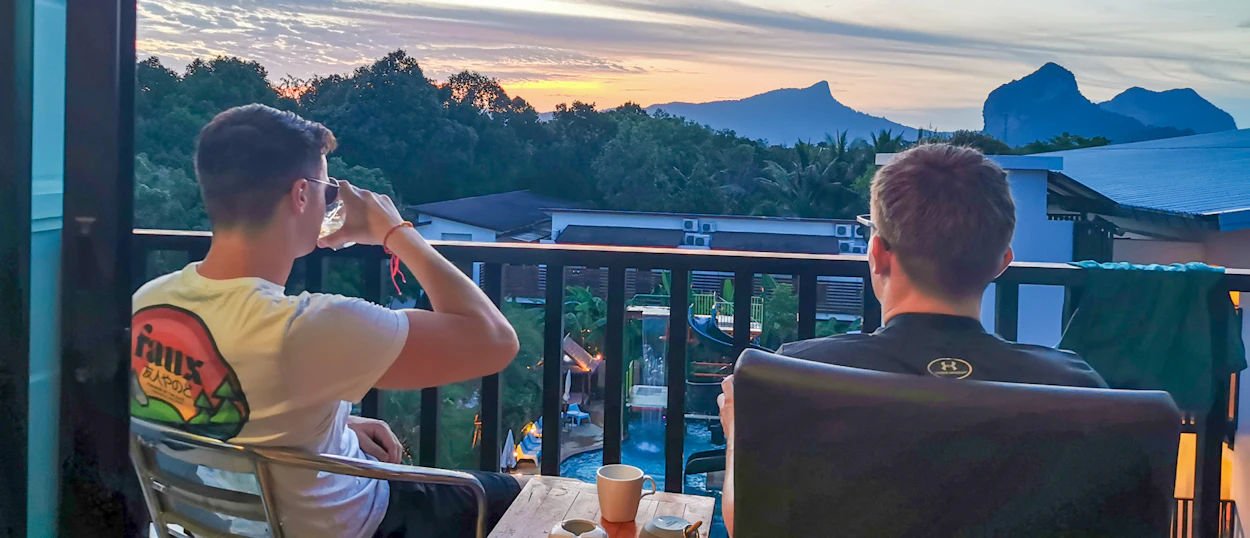 Two men relax on a balcony, watching a beautiful sunset over Krabi's dramatic limestone mountains.