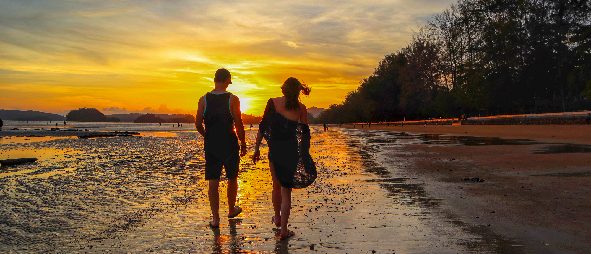 A couple walks along a wet beach at sunset in Krabi, Thailand, under a vibrant orange sky.