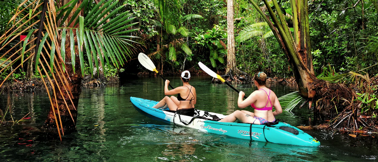 Two women paddle a blue kayak through a clear river in Krabi's lush mangrove jungle.