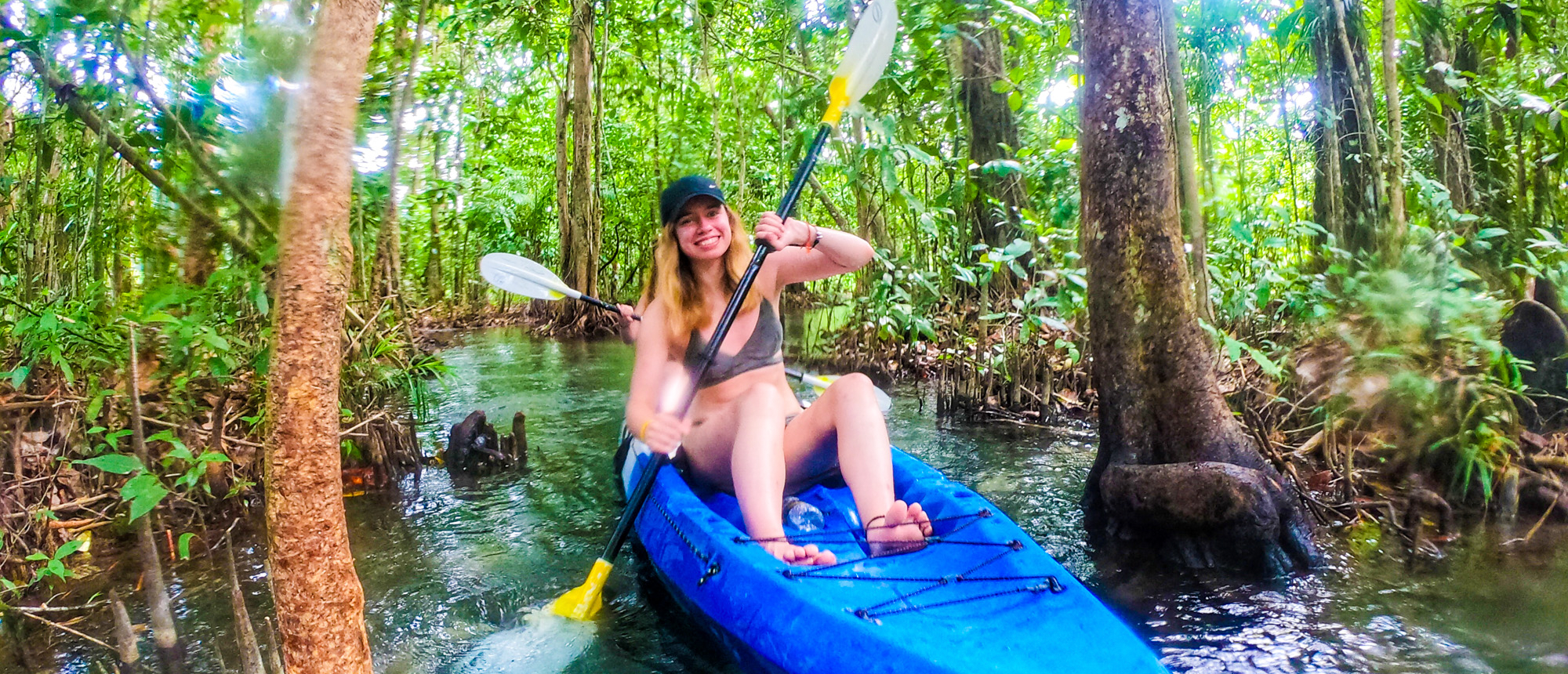 A smiling woman kayaks through a lush green mangrove forest in Krabi, Thailand.