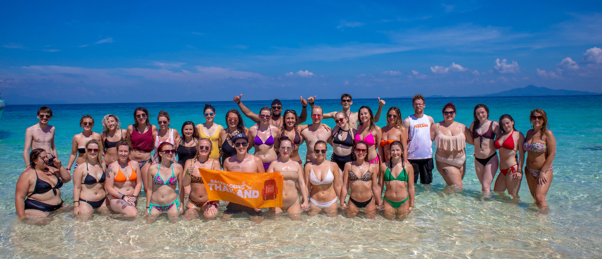 A joyful group of backpackers stands in the shallow, clear blue ocean water in Thailand.