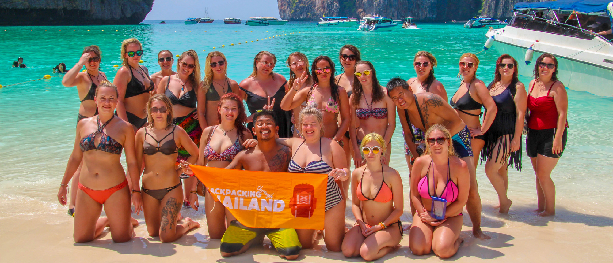 Smiling backpackers pose on the sandy beach of Koh Phi Phi, Thailand, with turquoise water and cliffs.