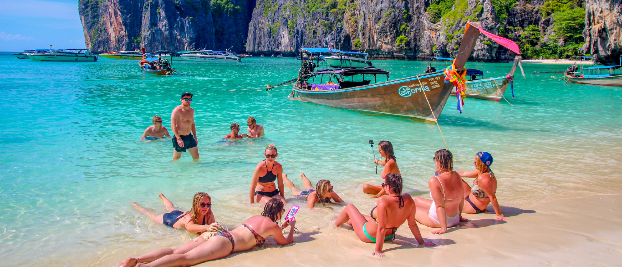 Happy travelers swim and relax on the sandy beach with longtail boats at Phi Phi Islands, Thailand.