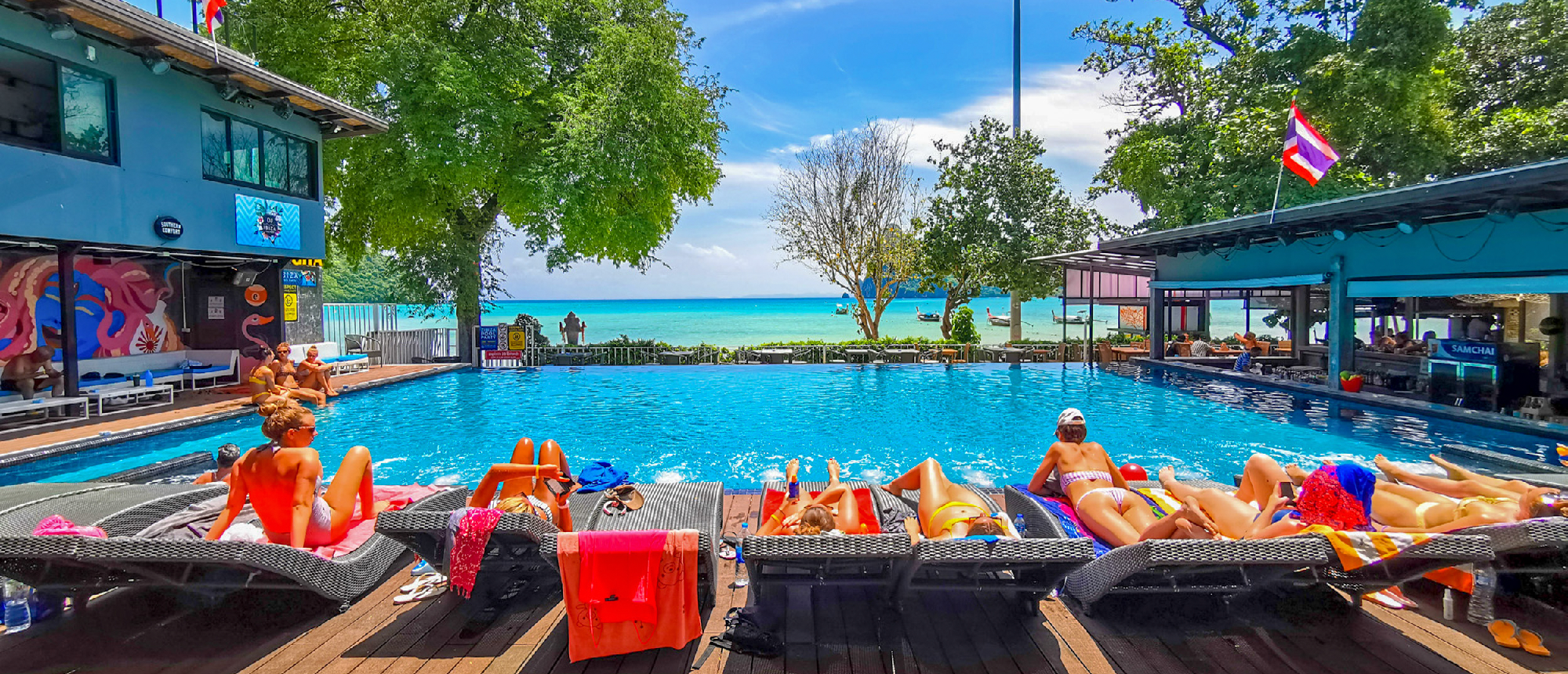 Sunbathers relax on lounge chairs by a vibrant blue pool overlooking the turquoise ocean in Thailand.