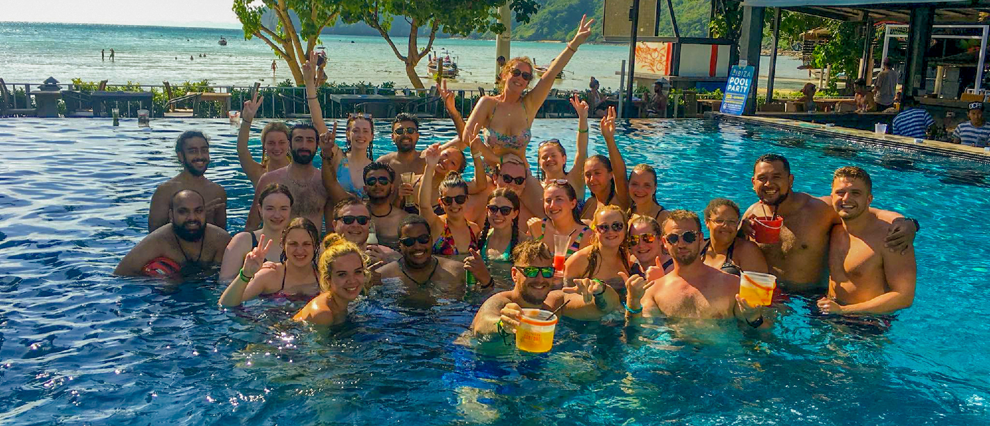 Happy backpackers in a sunny infinity pool with drinks, enjoying the ocean view on a Thai island.