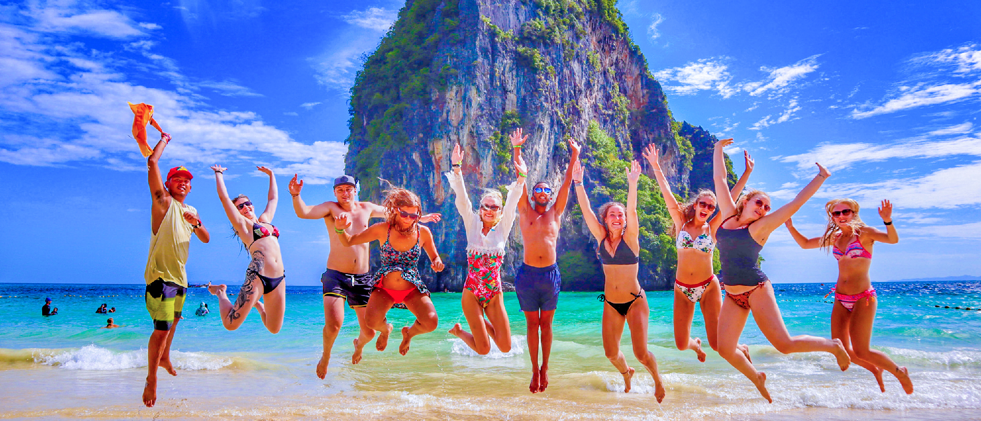 A joyful group of friends jumping on the white sands of Railay Beach, Thailand, with limestone cliffs.