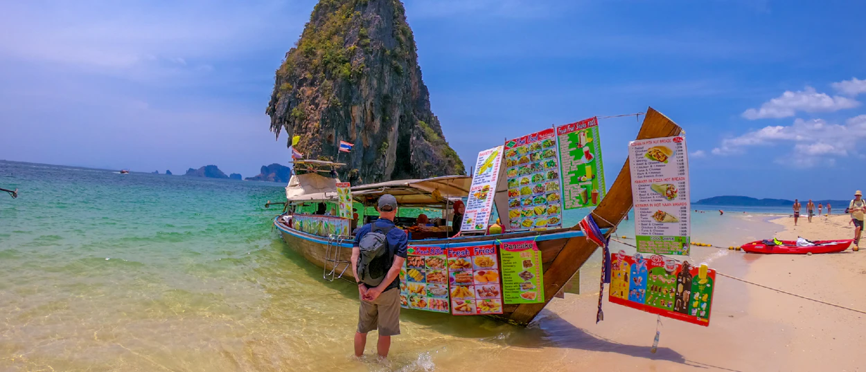 A man stands in clear water by a longtail boat on Railay Beach, Thailand, near limestone cliffs.
