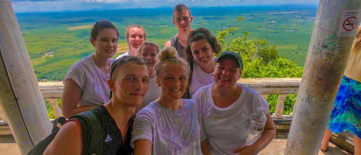A group of happy hikers smile at a viewpoint overlooking lush green landscapes in Krabi, Thailand.