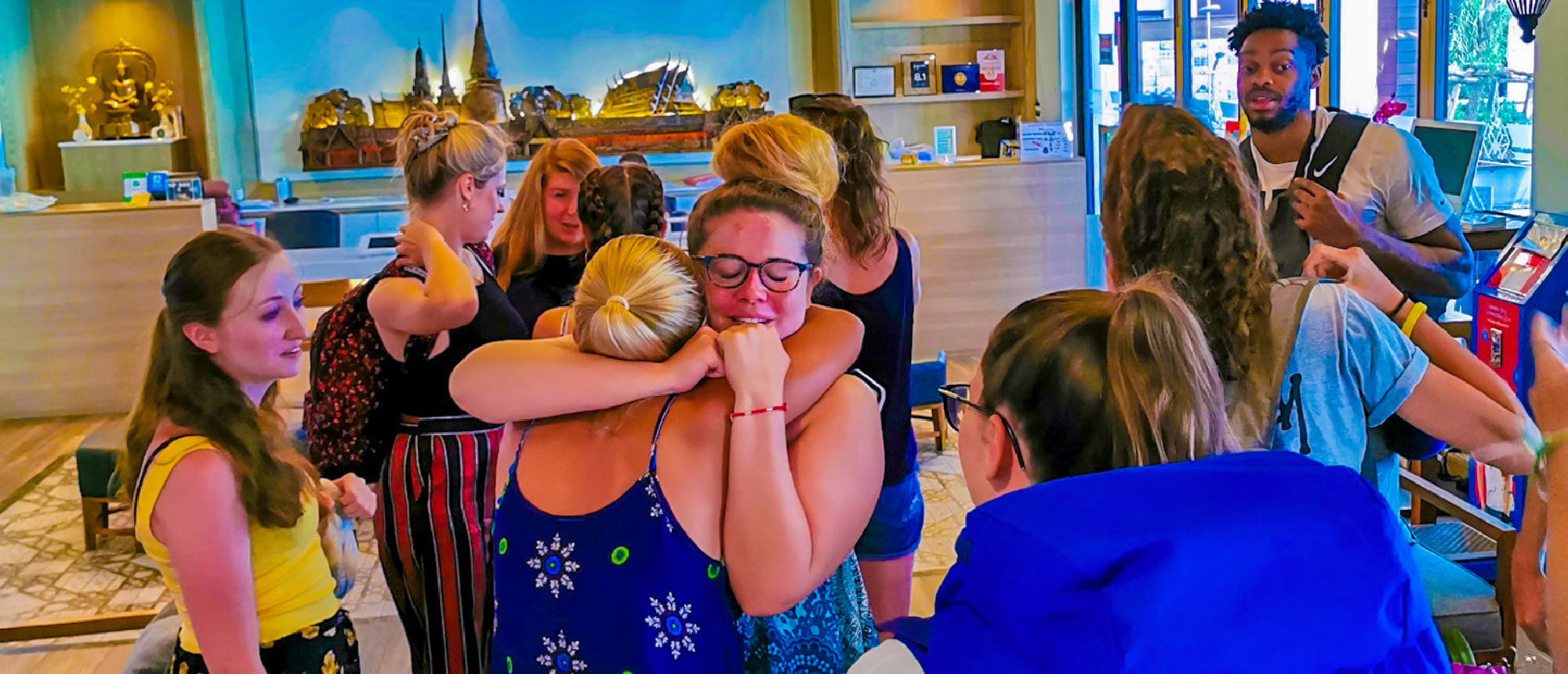 Emotional farewell as two women embrace in a hotel lobby, surrounded by their backpacking tour group in Thailand.