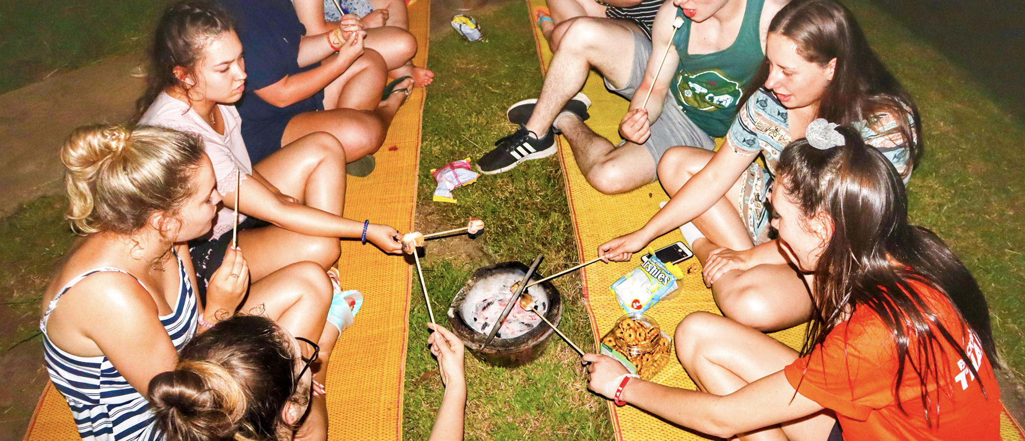 Young travelers gather around a campfire at night in Kanchanaburi, roasting marshmallows and enjoying fireside chats.