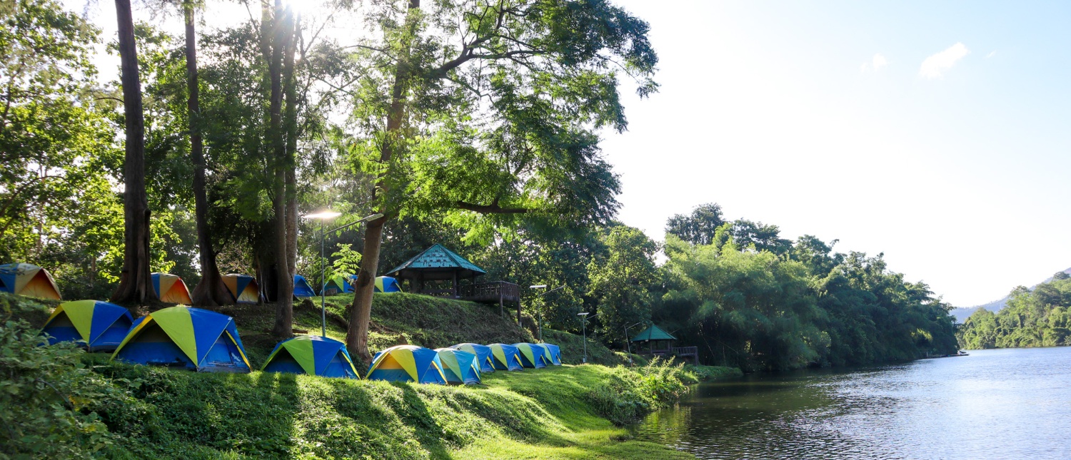 Colorful tents pitched along a lush, tree-lined riverbank in Kanchanaburi, Thailand, for serene camping.