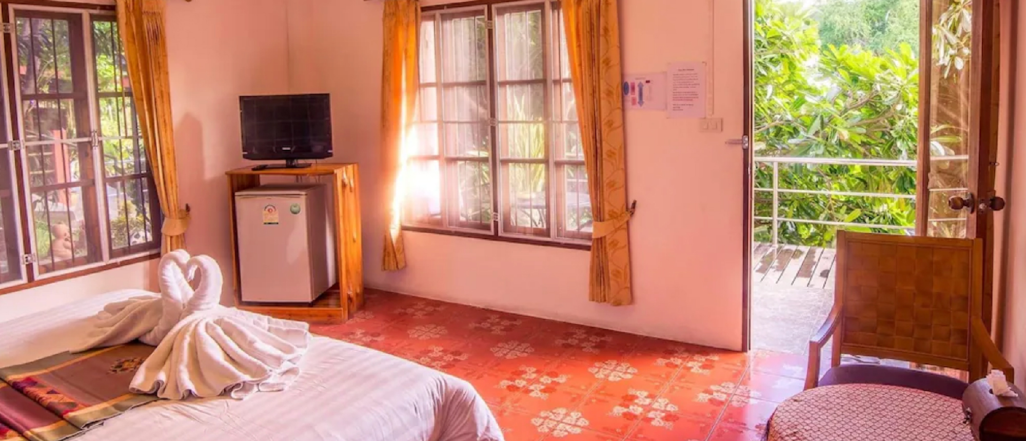 A bright hotel room with a bed, swan-folded towels, and a balcony view of lush greenery.