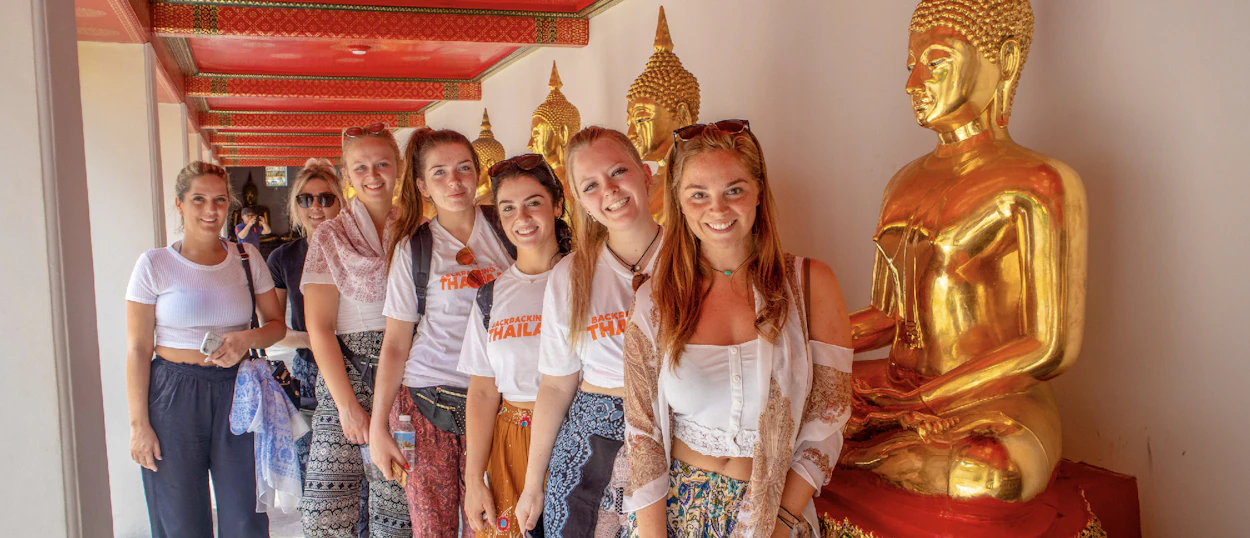 Seven smiling women pose in a temple hallway lined with golden Buddha statues in Bangkok.