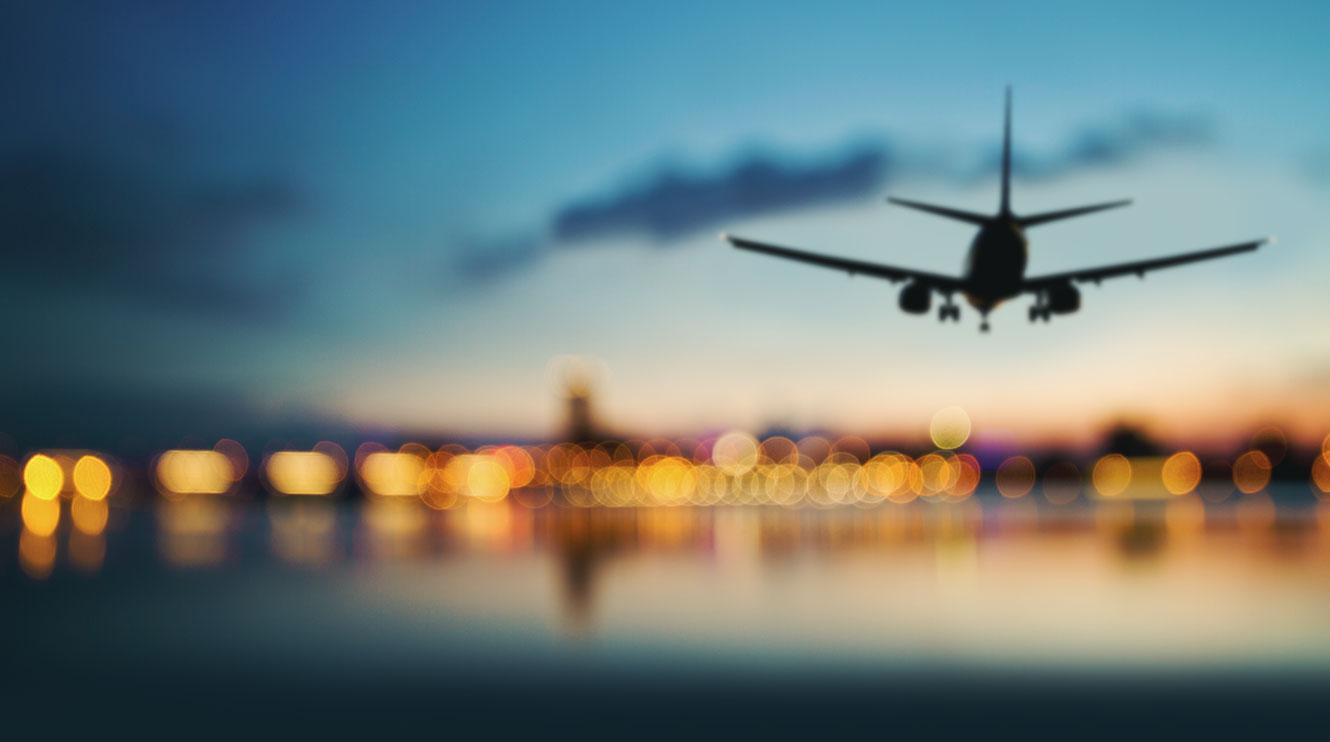 An airplane silhouette against a colorful twilight sky, with blurred city lights reflecting below.