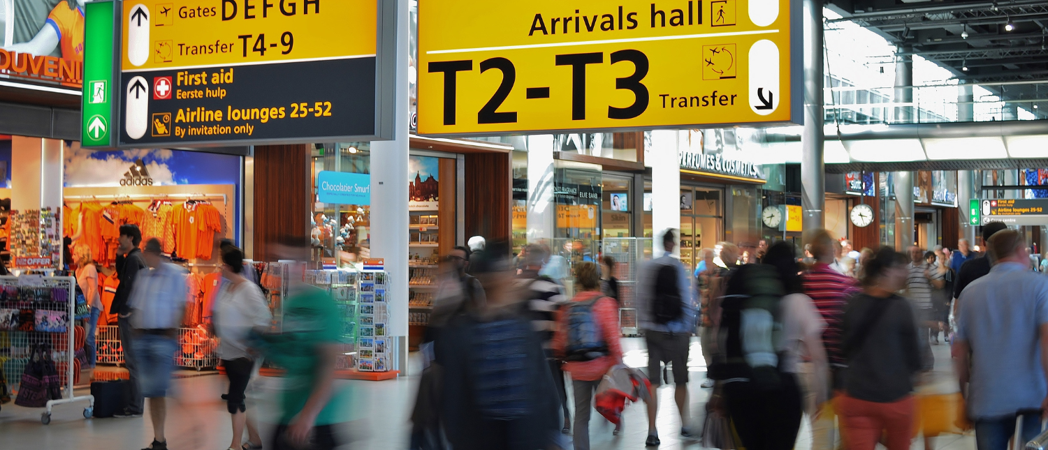 Travelers with backpacks move through a busy airport terminal with yellow directional signs.