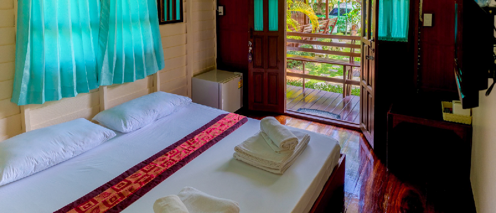 A welcoming homestay bedroom in Amphawa, Thailand, with a bed, towels, and an open door to nature.