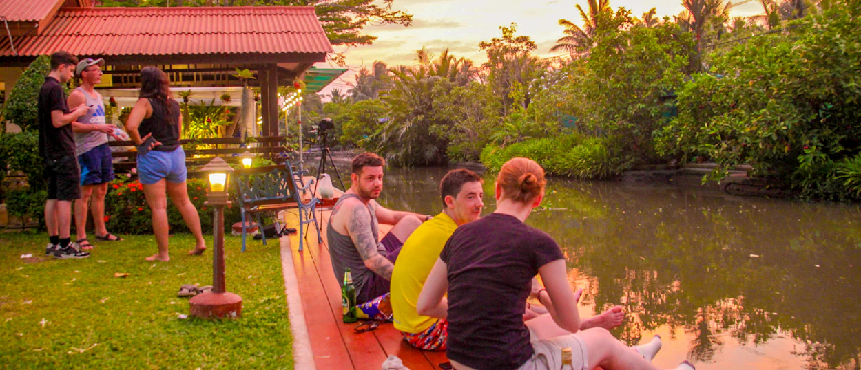 Travelers socialize by a canal at sunset in Amphawa, Thailand, enjoying a relaxed homestay evening.