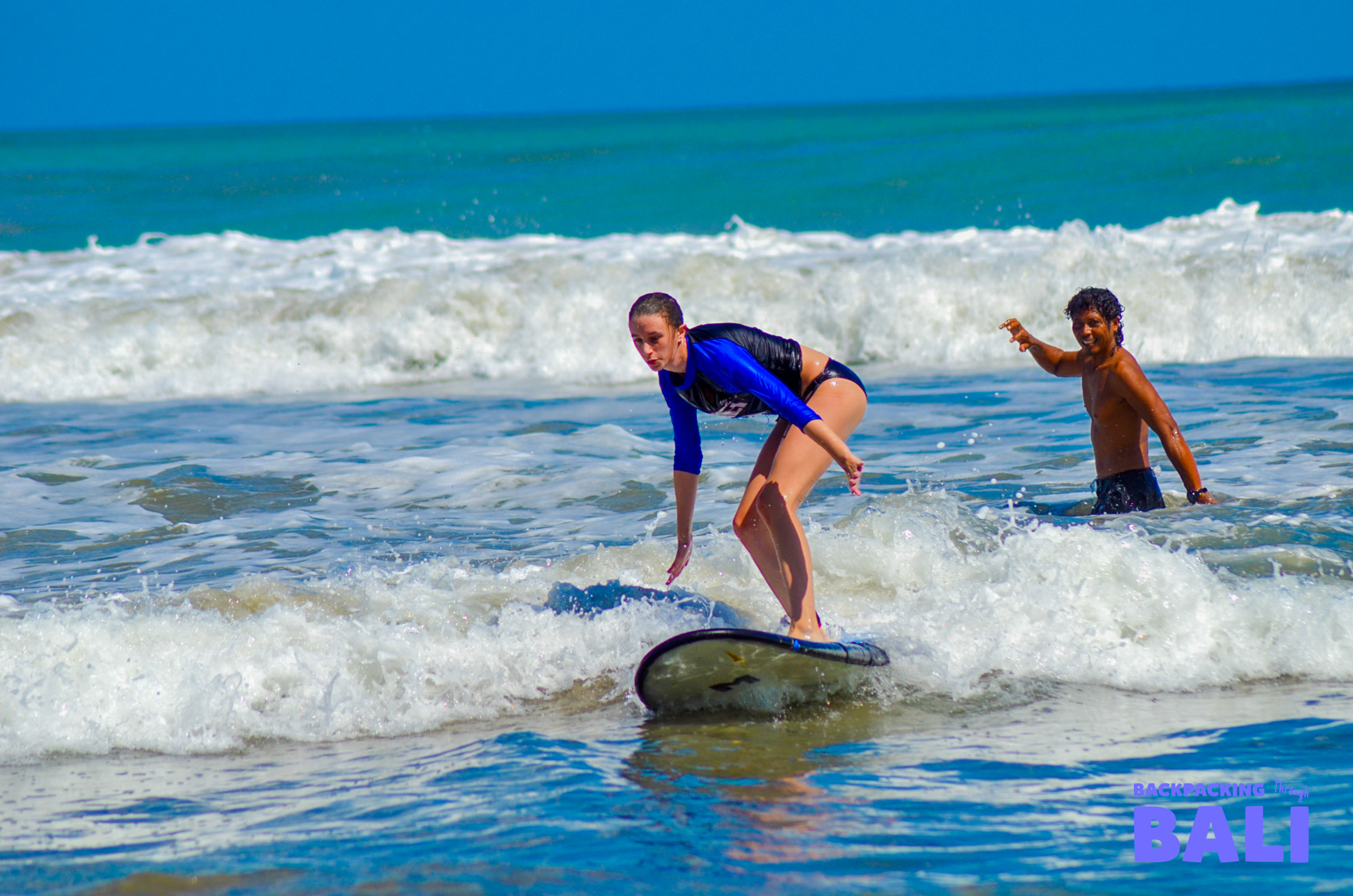 A woman surfs a small wave with a man watching in the sunny Bali ocean.