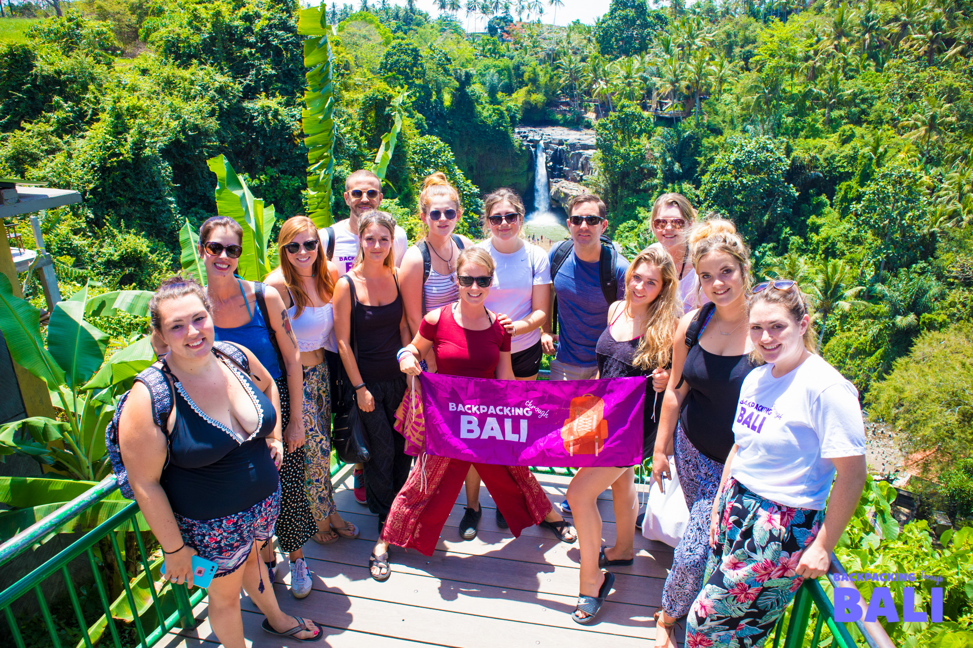 A group of smiling backpackers holding a Bali banner in front of a tropical waterfall.