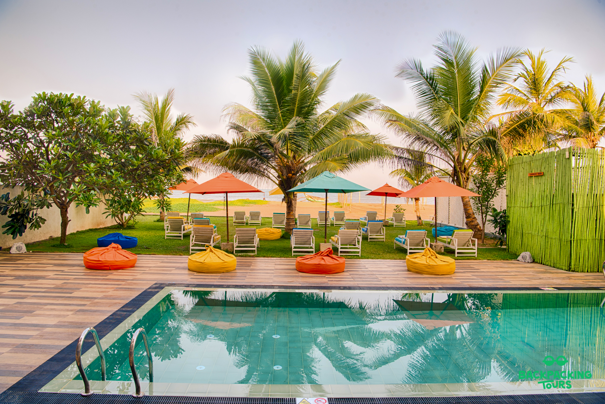 A vibrant resort pool area with colorful beanbags, sun loungers, palm trees, and a beach in Colombo.