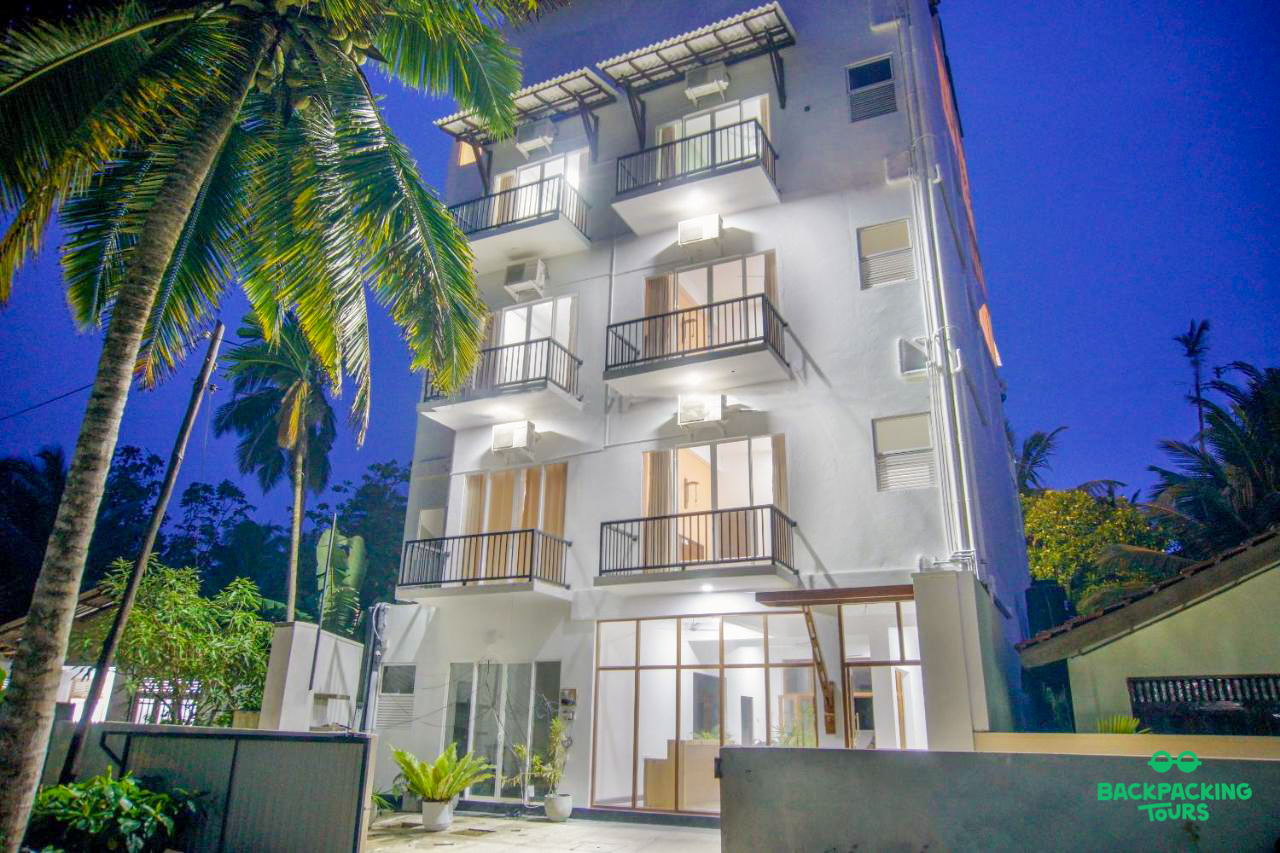 The bright white accommodation building with balconies and a palm tree at night in Weligama.