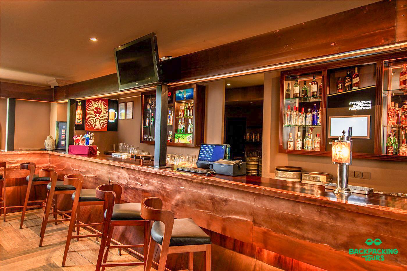 A stylish, empty bar featuring a polished wooden counter, comfortable stools, and backlit liquor shelves.