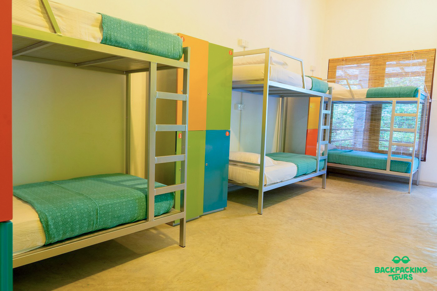 A spacious and clean hostel dorm room featuring multiple bunk beds with teal bedding and colorful lockers.