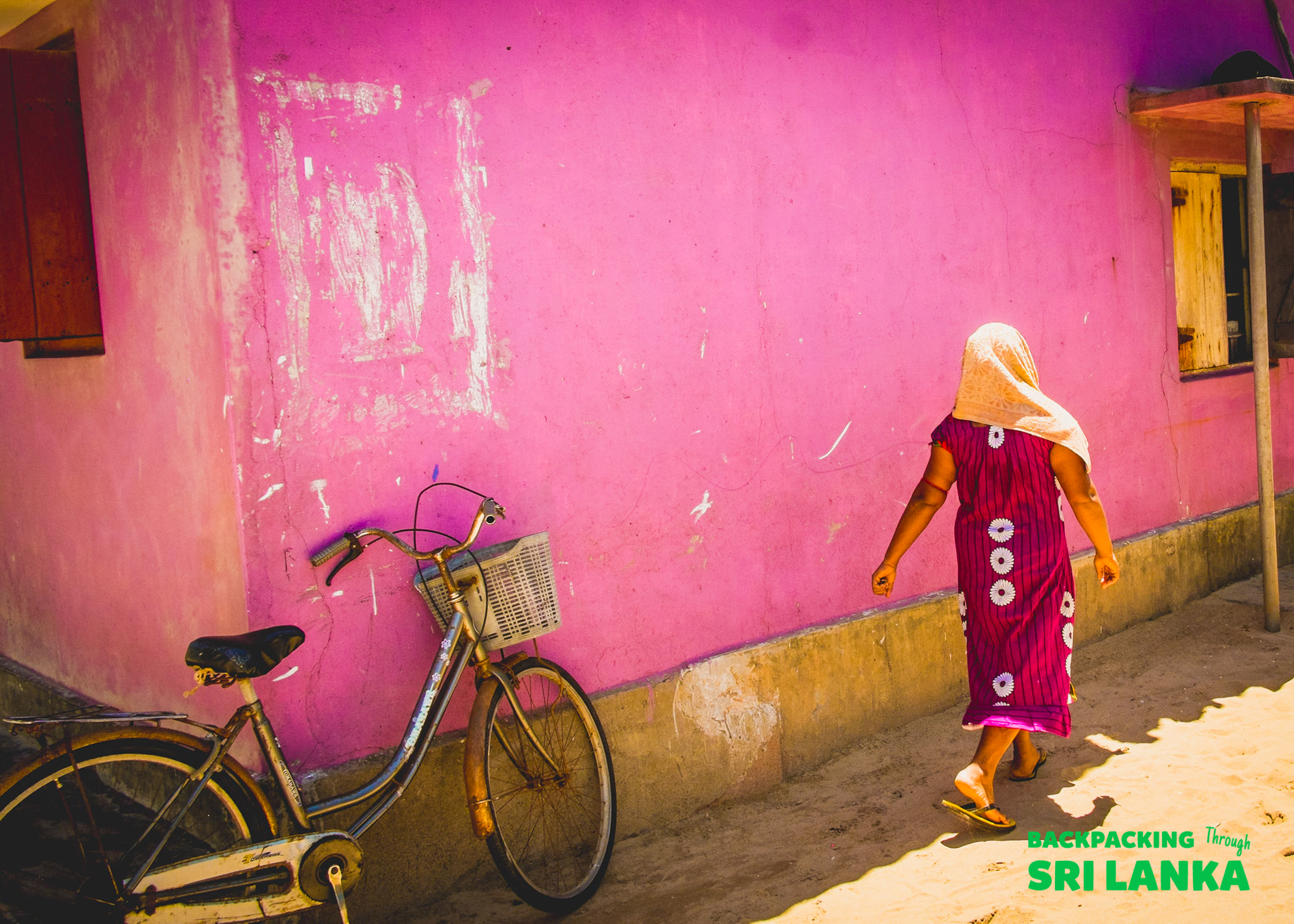 A woman walks past a bicycle and a bright pink building wall in Colombo.