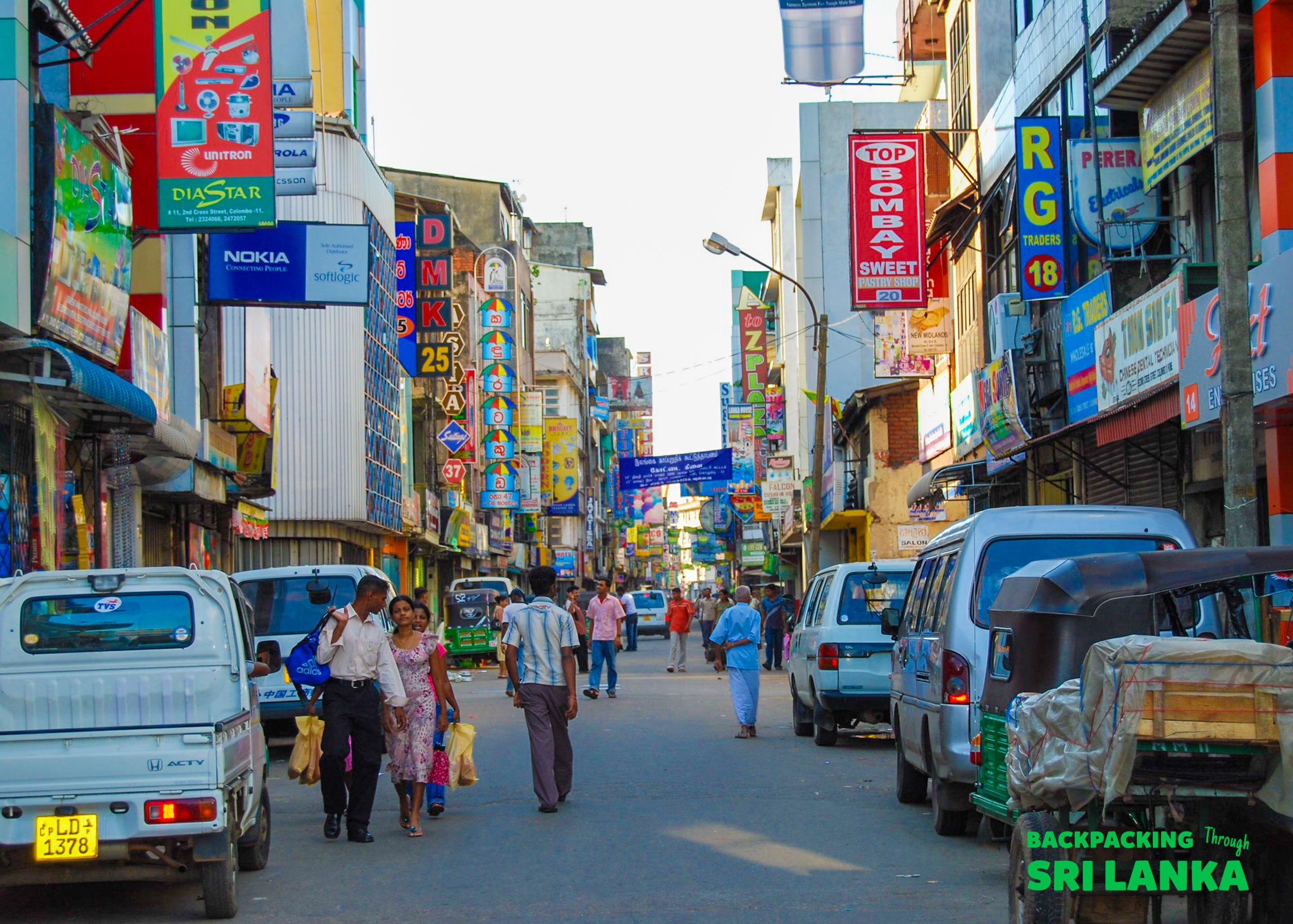 People and vehicles fill a vibrant street in Colombo, Sri Lanka, lined with glowing signs.