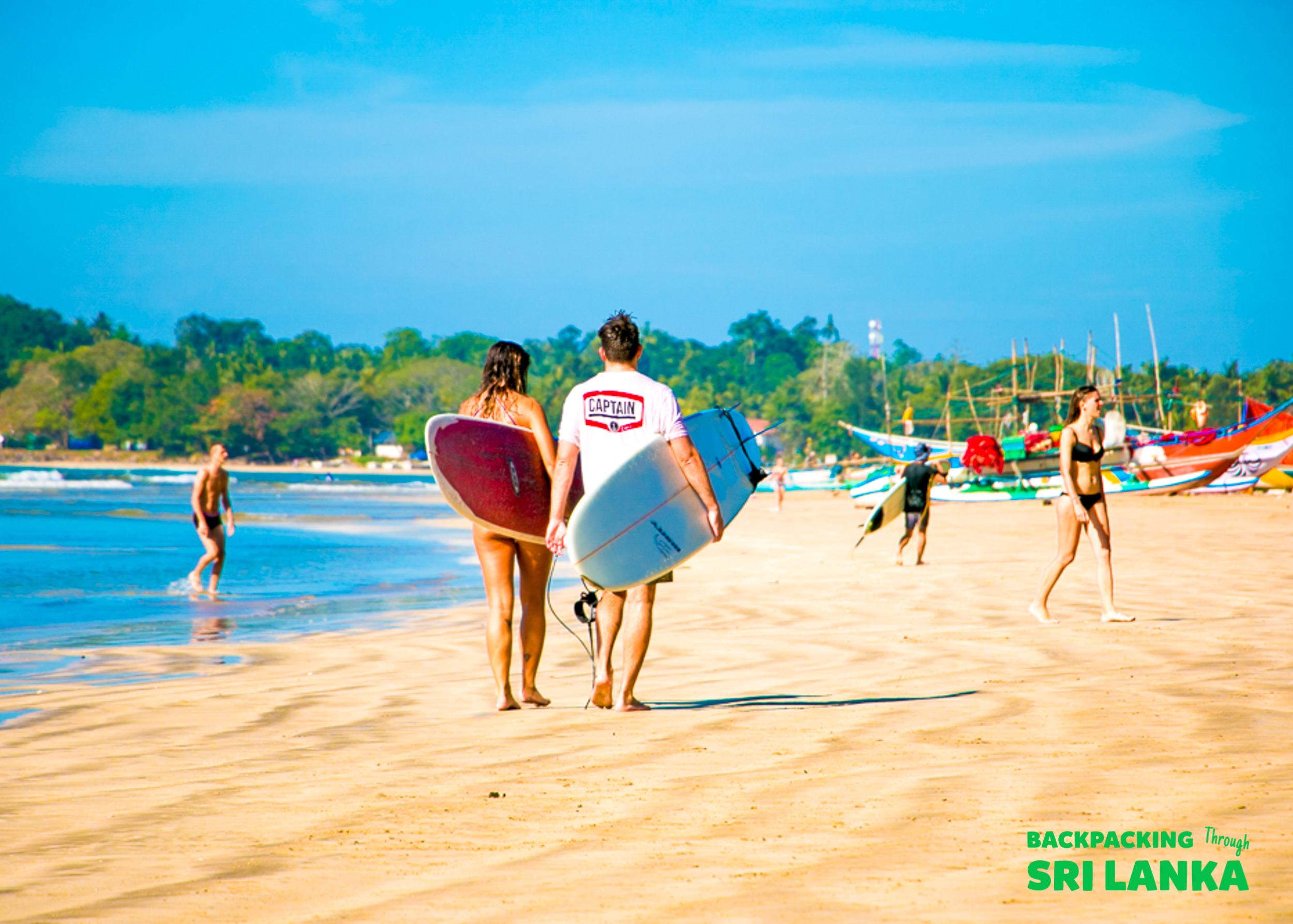 Two people carrying surfboards walk along the sunny Weligama beach in Sri Lanka.