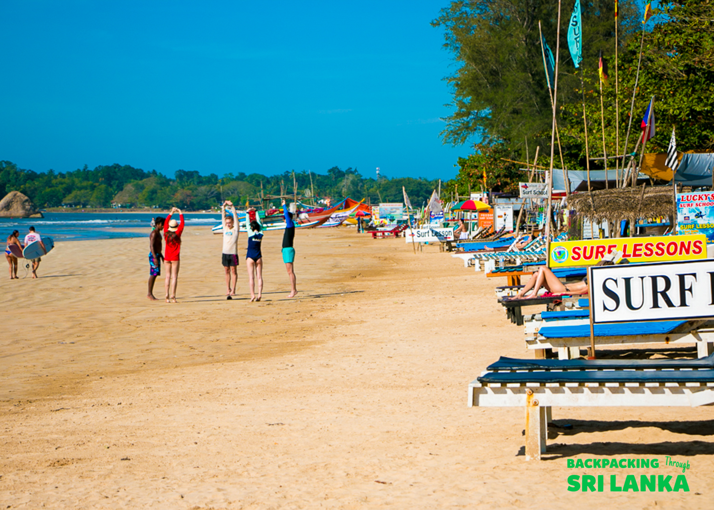 Beginners stretch on Weligama Beach, Sri Lanka, before a surf lesson under a clear blue sky.