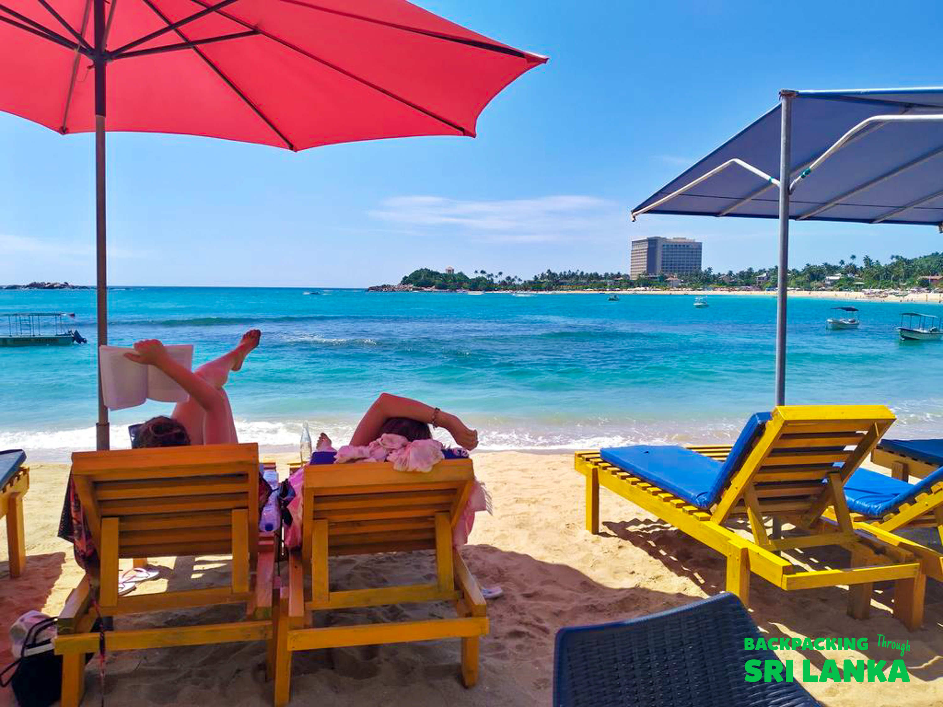 Two people relax on sun loungers under colorful umbrellas on a sunny beach in Unawatuna.