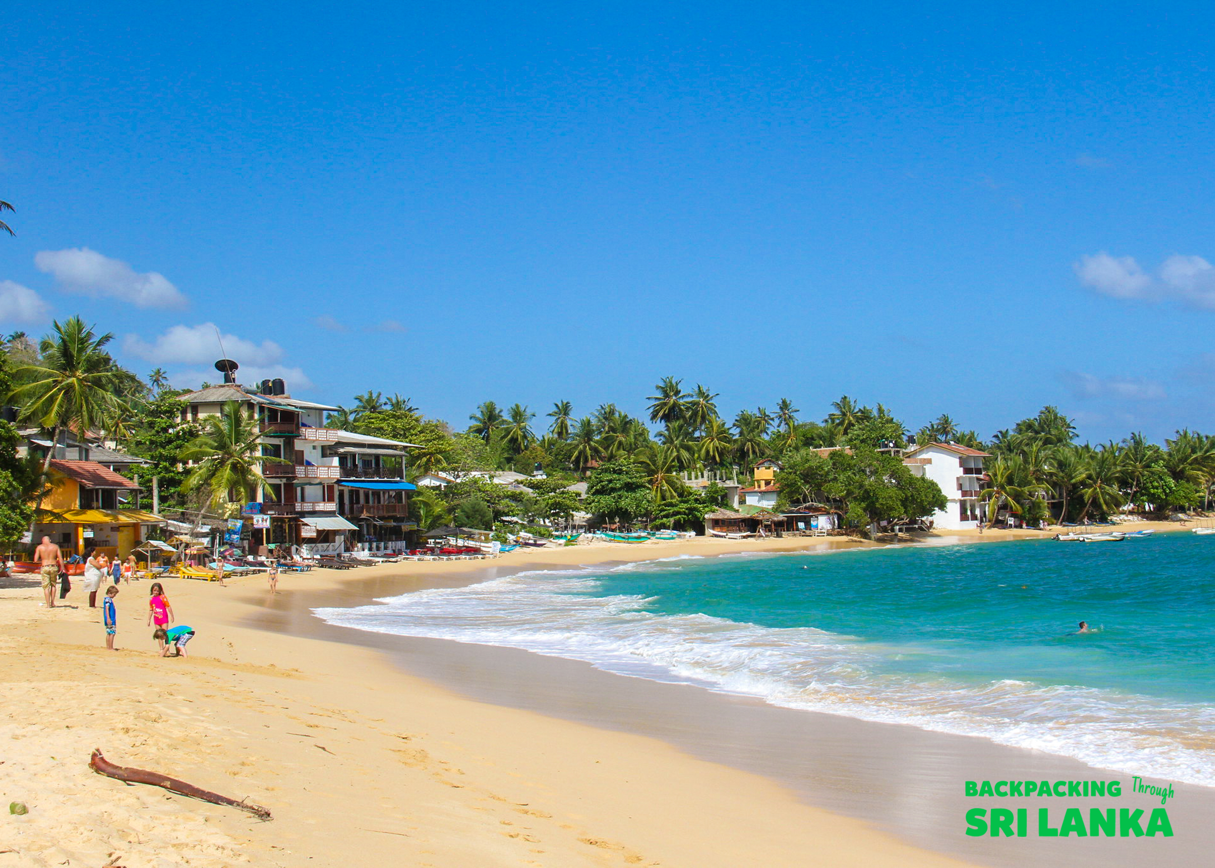 Tourists walk along the beautiful sandy beach of Unawatuna with clear blue water and palm trees.