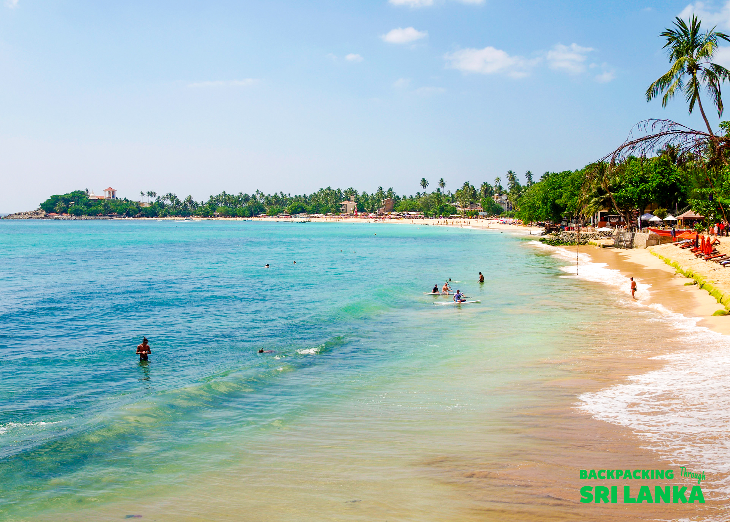 People swim and enjoy the clear turquoise waters of a sunny tropical beach in Unawatuna, Sri Lanka.