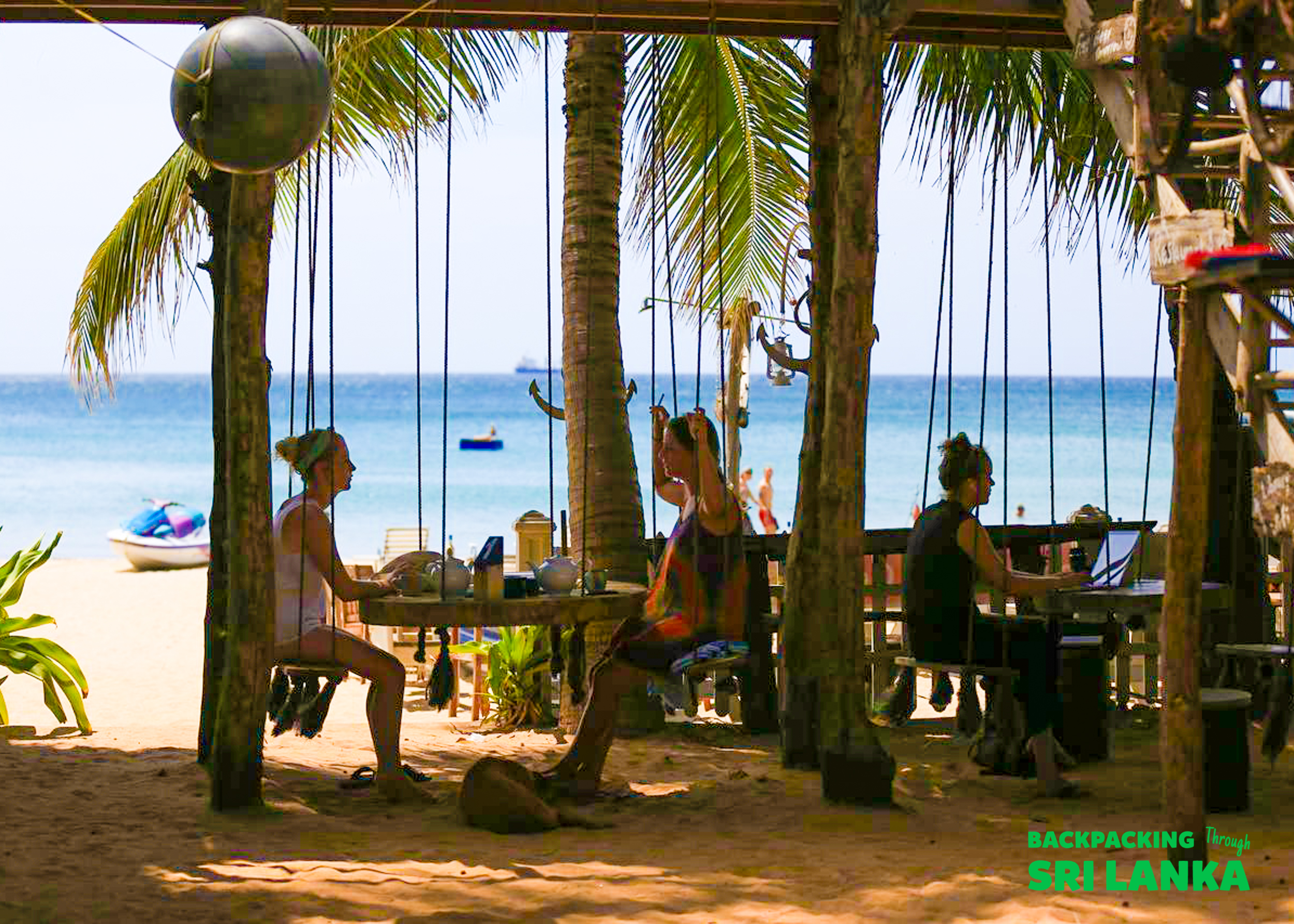 Three travelers enjoy drinks and work at a beachside bar with swing seats in Unawatuna, Sri Lanka.
