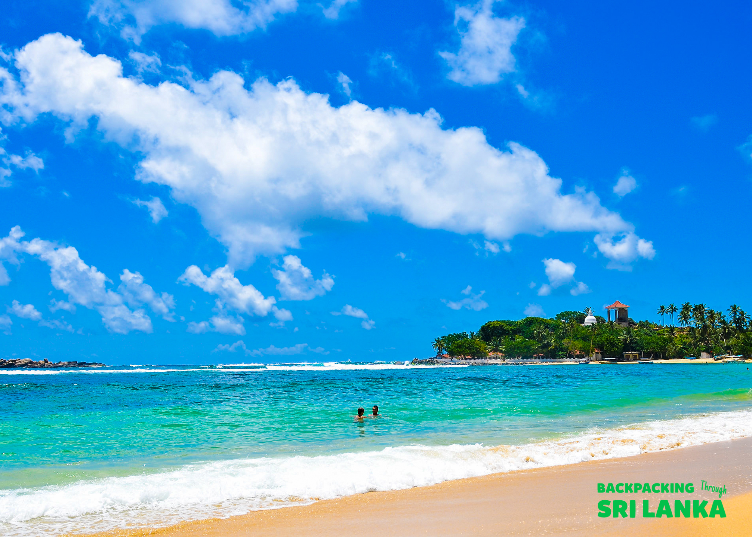 Two people swim in the clear turquoise waters of Unawatuna Beach under a bright blue sky.