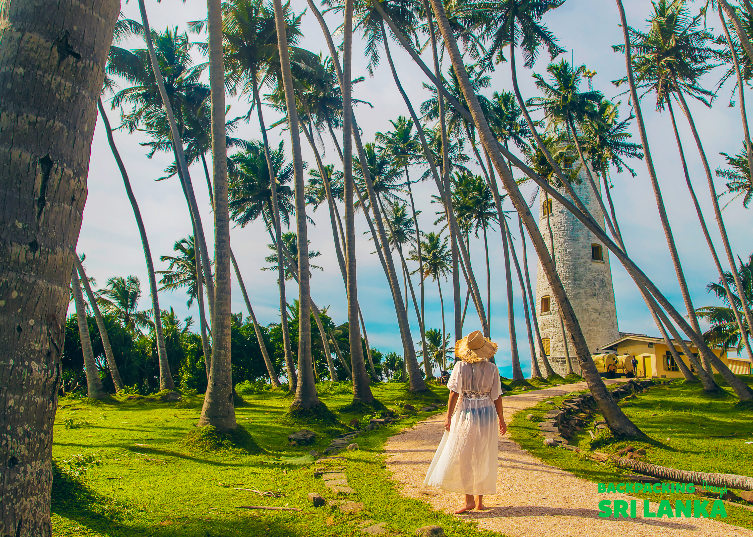 A woman in a white dress walks towards the historic Galle Lighthouse surrounded by palm trees.