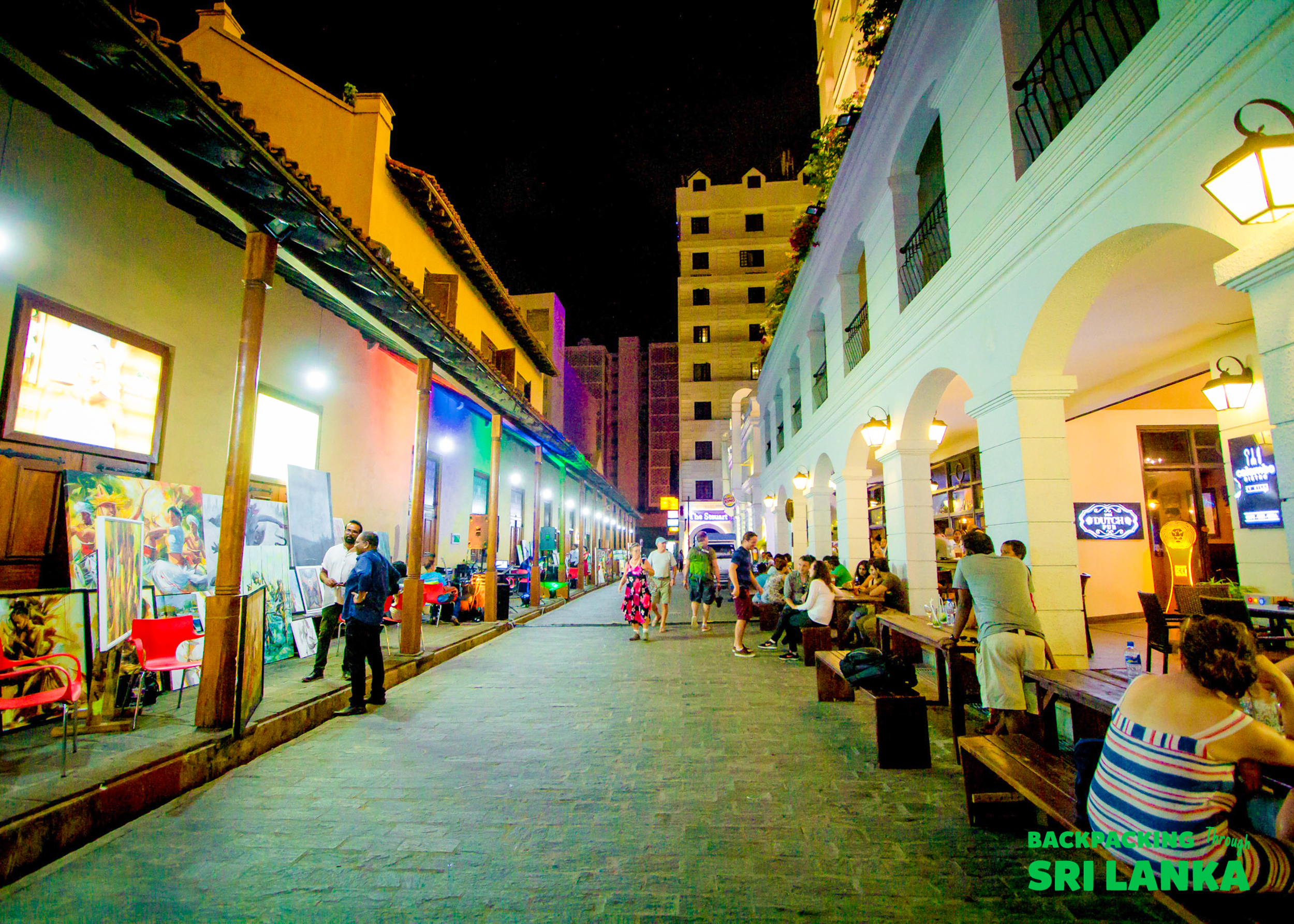 A vibrant night market street in Colombo with people dining outdoors and browsing colorful art stalls.