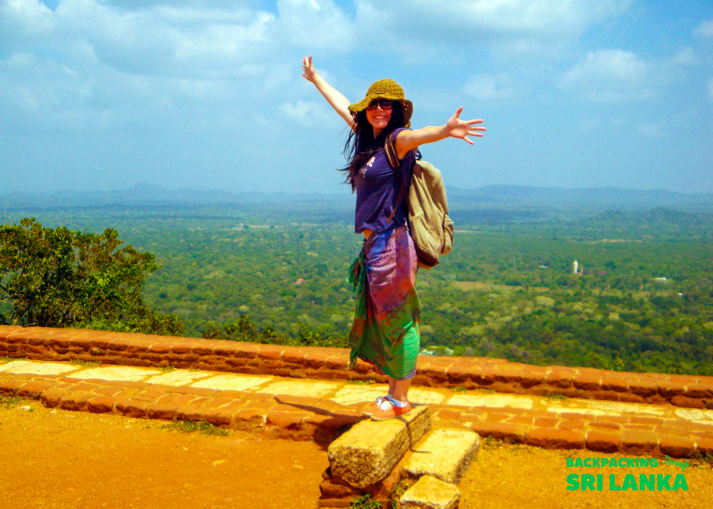 A joyful backpacker stands on Sigiriya Rock, arms wide, overlooking the lush green landscape of Sri Lanka.