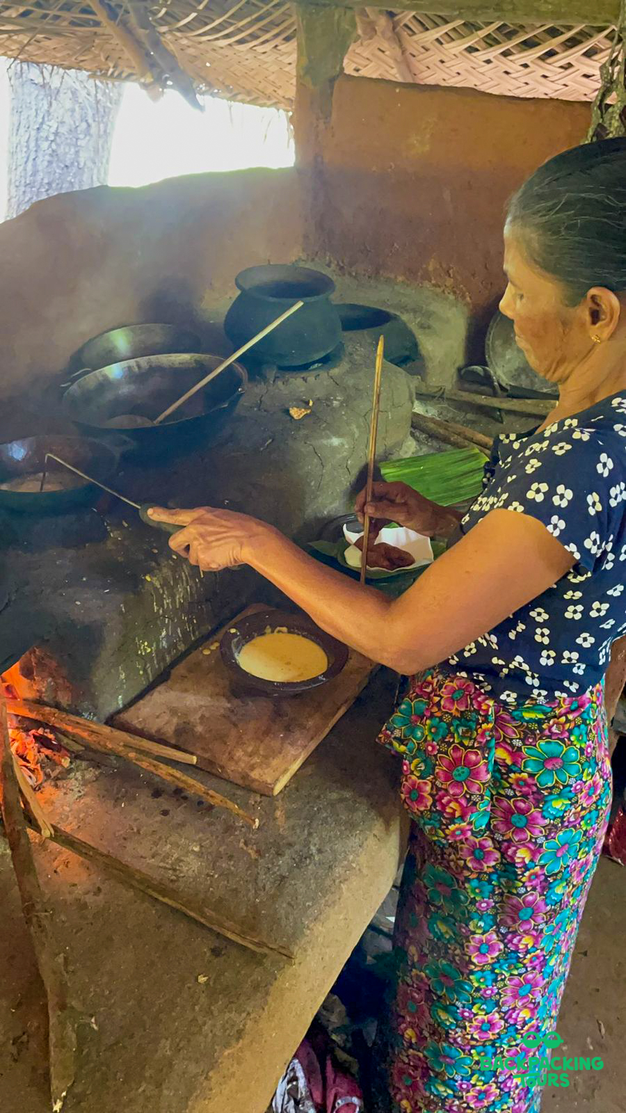 A woman cooks traditional food over an open fire in a rustic Sri Lankan village kitchen.