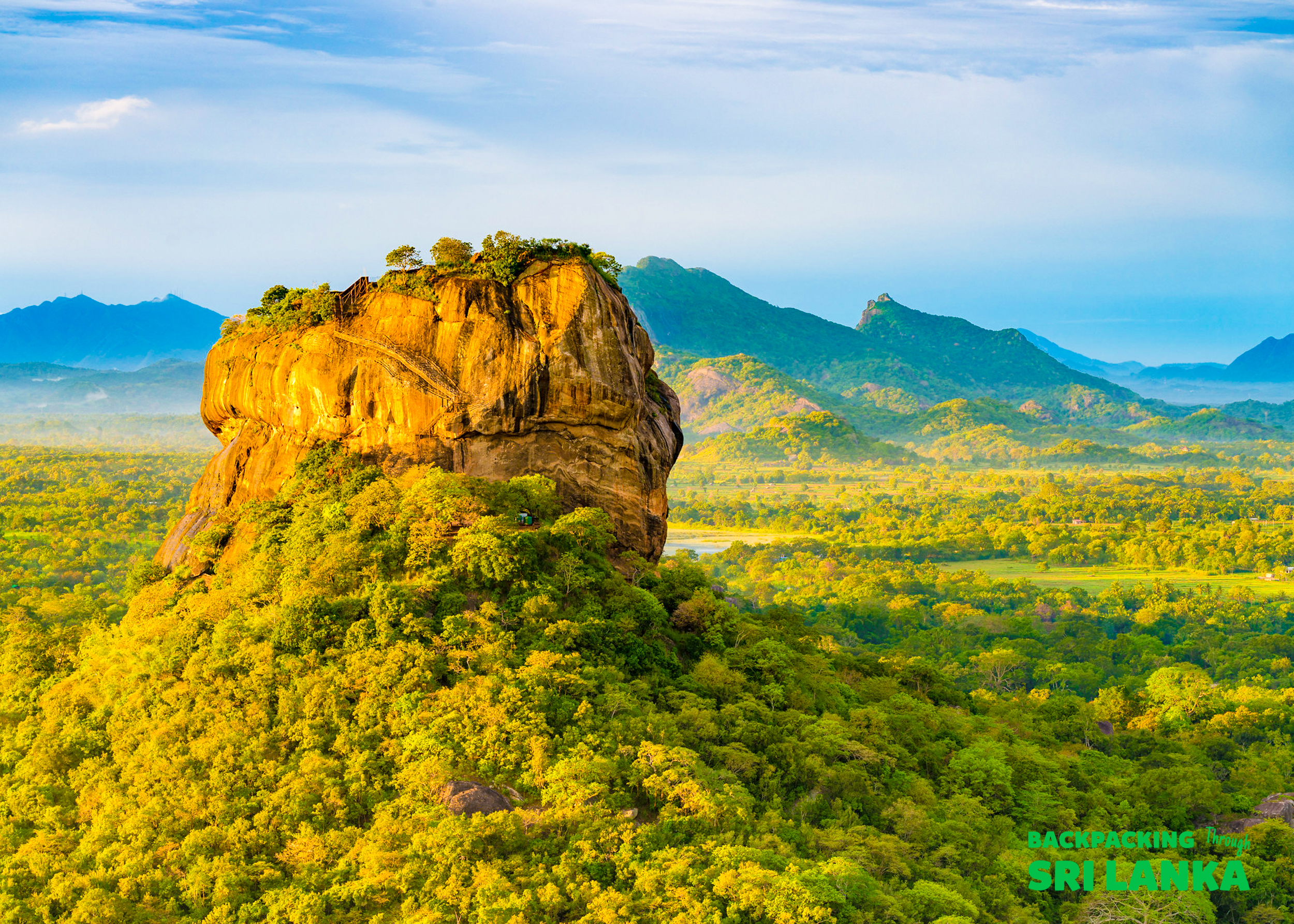 Sigiriya Rock, a majestic golden monolith, rises above a lush green forest under a clear blue sky.
