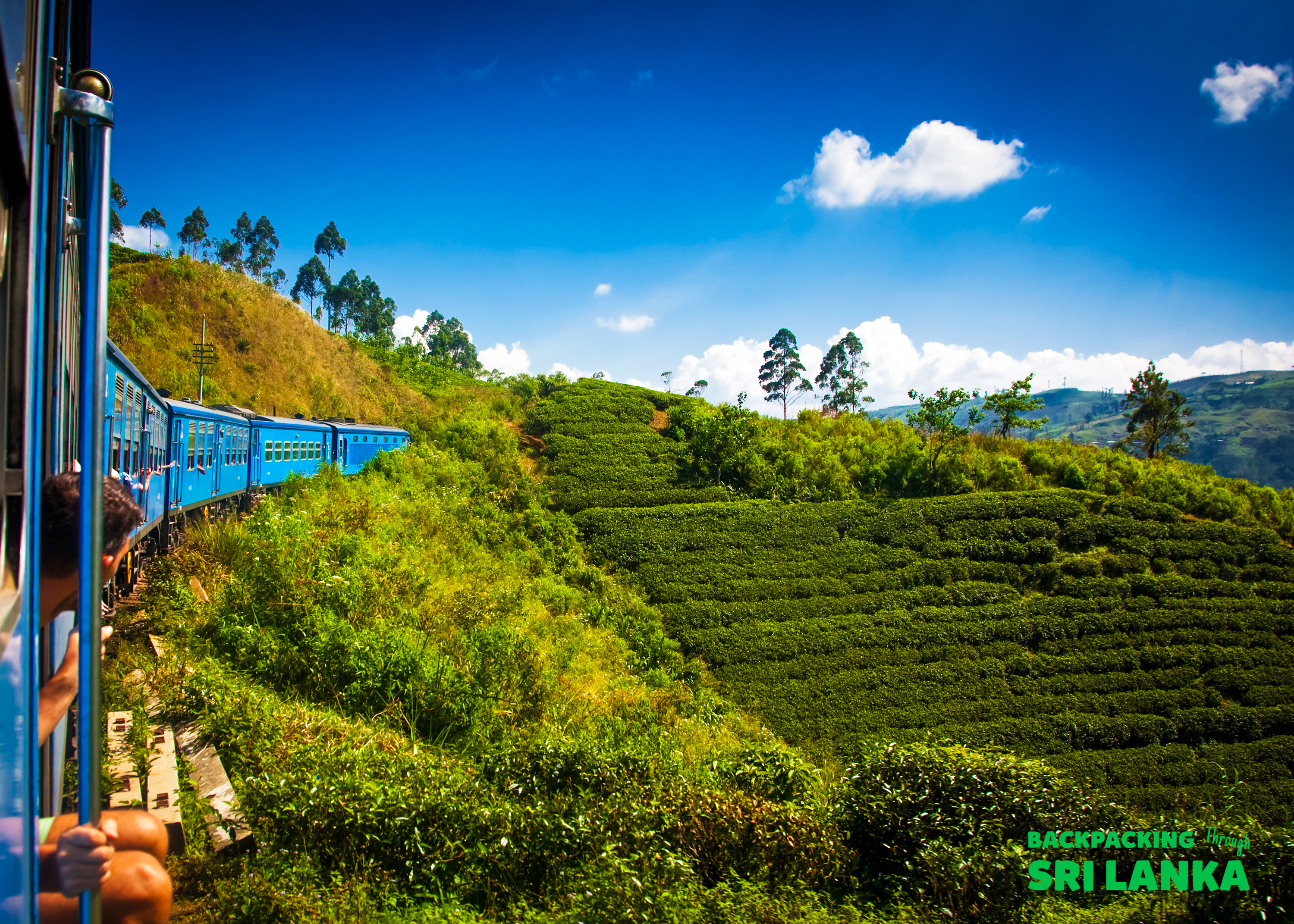 A blue train winds through vibrant green tea plantations under a bright sky in Ella, Sri Lanka.