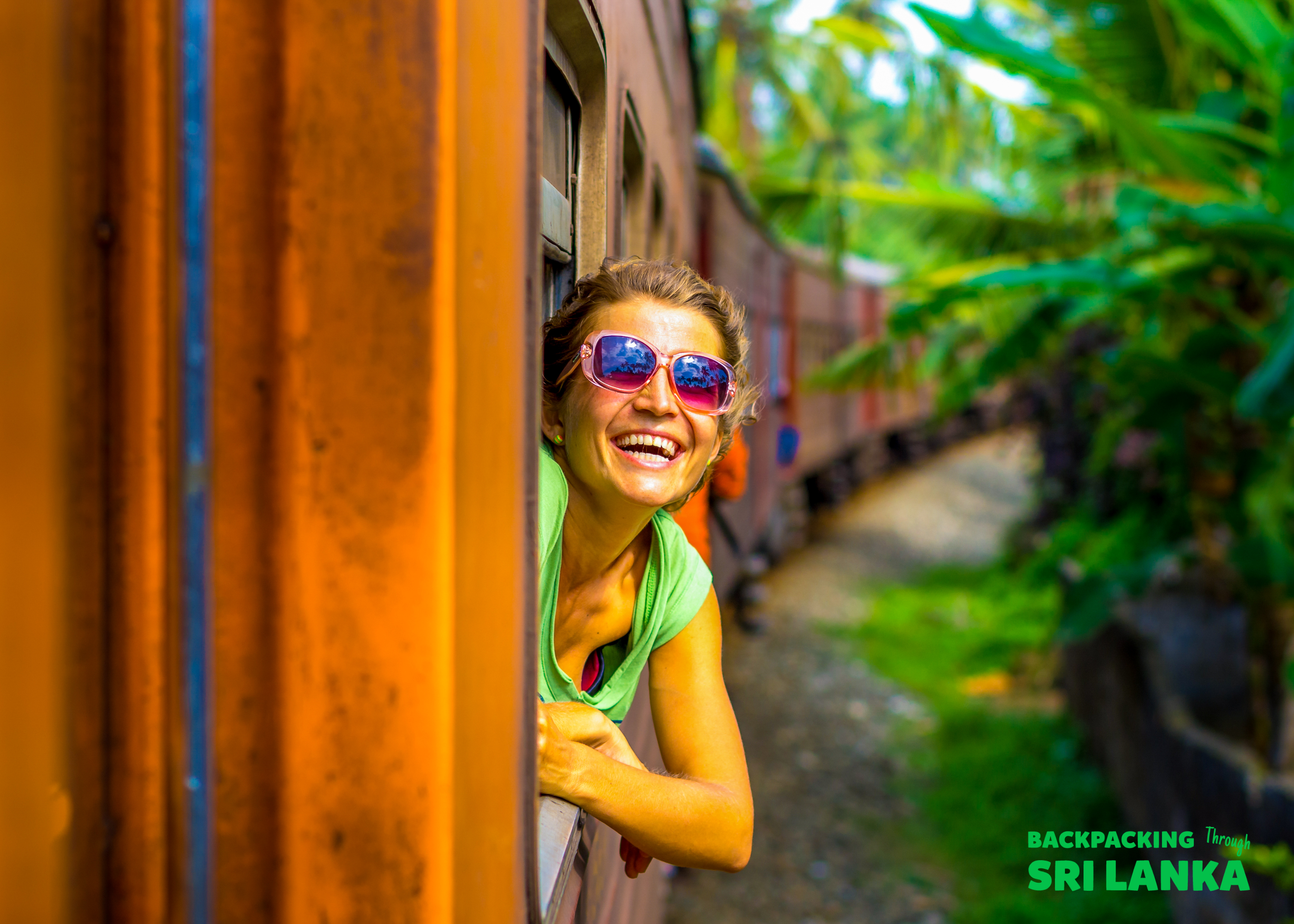 A smiling woman with sunglasses leans out of a train window, enjoying the scenic ride through Sri Lanka.