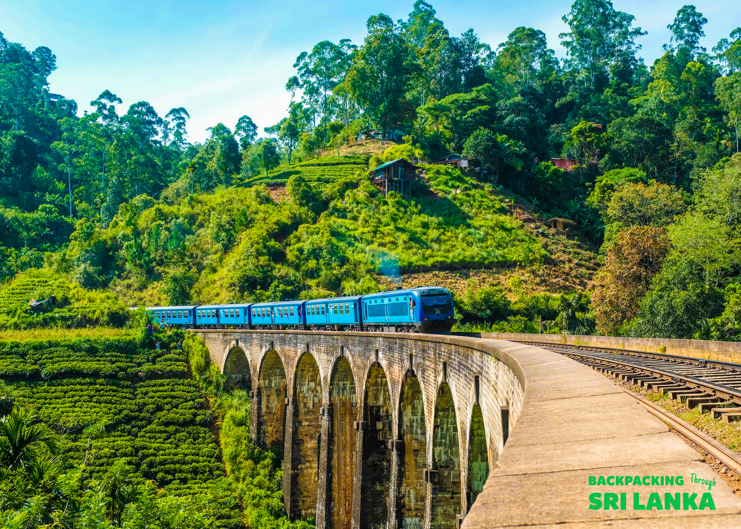 A blue train crosses the iconic Nine Arch Bridge surrounded by lush green tea plantations in Ella, Sri Lanka.